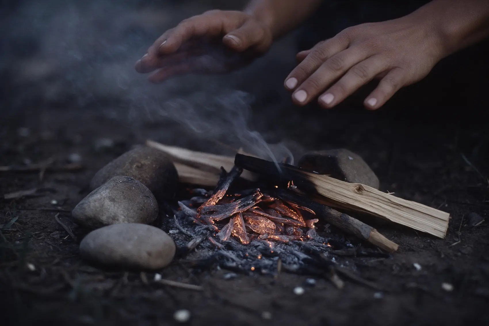 Kleines Ritualfeuer auf blanker Erde zur blauen Stunde, zwei Hände nahe der Glut.